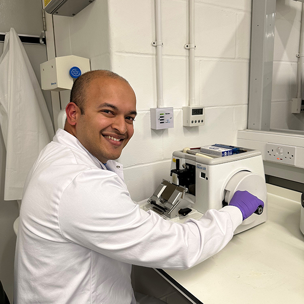 A photo of Dr. Avirup Chowdhury sitting at a machine in his research lab, looking back over his right shoulder and smiling.